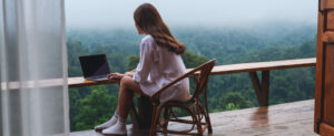 A woman sitting in her balcony looking at her laptop with a view of greenery in the distance